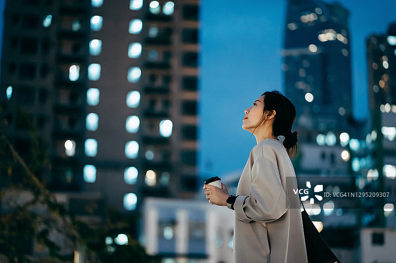 沉思的年轻亚洲女人，喝着咖啡，看着天空，而站在高层照明的城市建筑在晚上图片素材