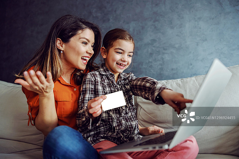 微笑的母女坐在沙发上，用笔记本电脑在网上购物。女儿拿着信用卡，指着她想买的笔记本电脑上的玩具。图片素材