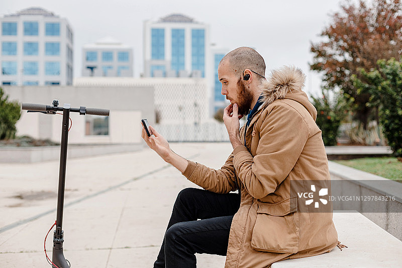 一个戴着蓝牙耳机的年轻人坐在长椅上，在一辆电动车旁边进行视频通话。图片素材