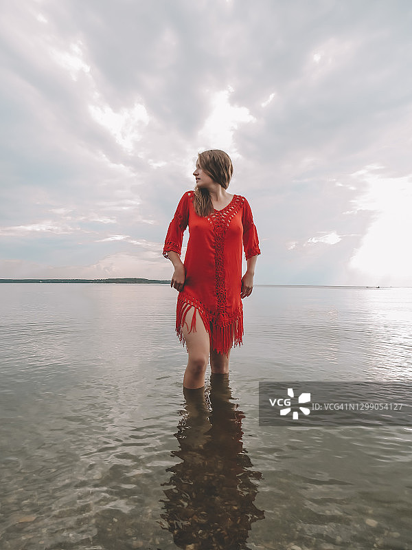 一位漂亮的年轻女子，一头金色长发，穿着红色沙滩裙，身材苗条。她走在海湾浅滩的水面上图片素材