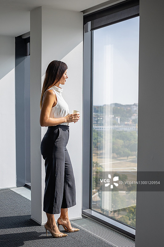 优雅的女商人站在办公室的大窗户旁喝咖啡休息图片素材