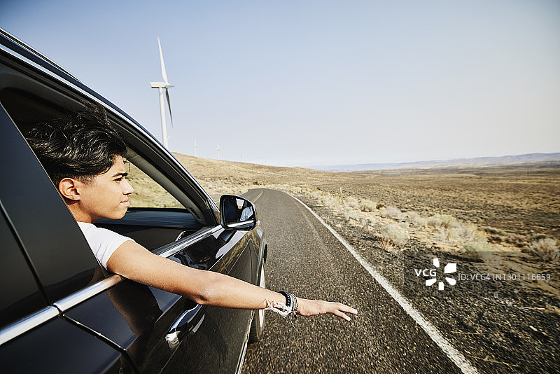 在沙漠里，一个十几岁的男孩把手臂伸出车窗，看着风力涡轮机图片素材