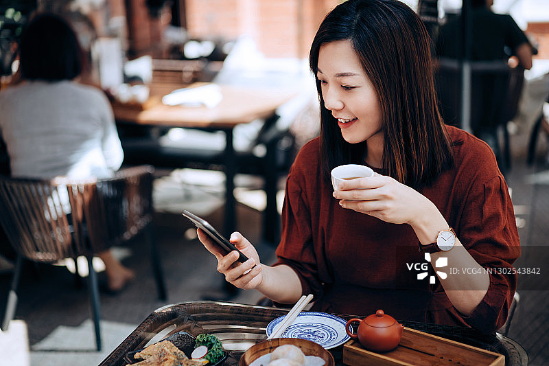 美丽微笑的年轻亚洲女子使用智能手机，同时享受新鲜供应的传统点心与中国茶在一个户外中国餐厅。传统的中国文化，美味茶，外出就餐的生活方式图片素材