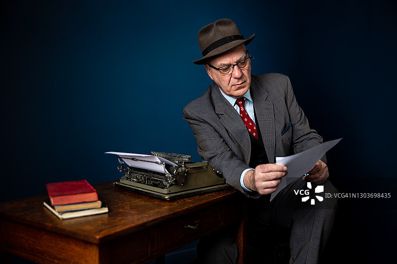 成熟的成年作家穿着西装和帽子坐在一个木制的书桌与老式打字机，阅读一页从故事或文章他写，复制空间图片素材