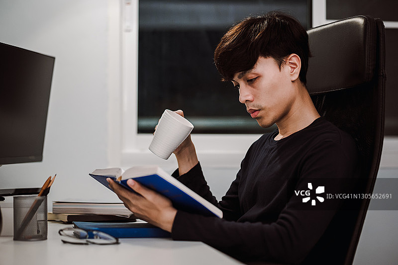 年轻帅气的亚洲男人在工作桌前看书喝茶，学习知识和概念图片素材