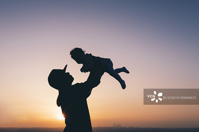 在美丽的日落中，一个妈妈用帽子把她的孩子举在空中的剪影。图片素材
