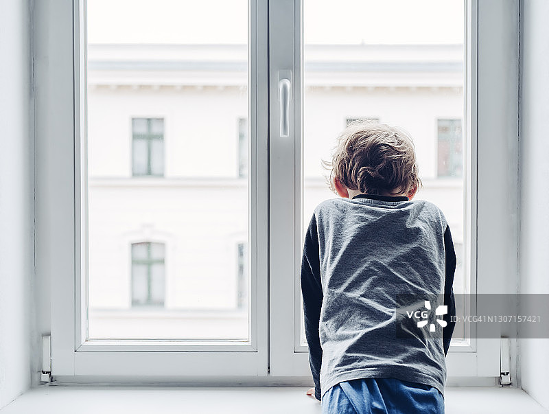 悲伤的小男孩望着窗外。在封锁或隔离期间，他不允许会见或探望朋友图片素材