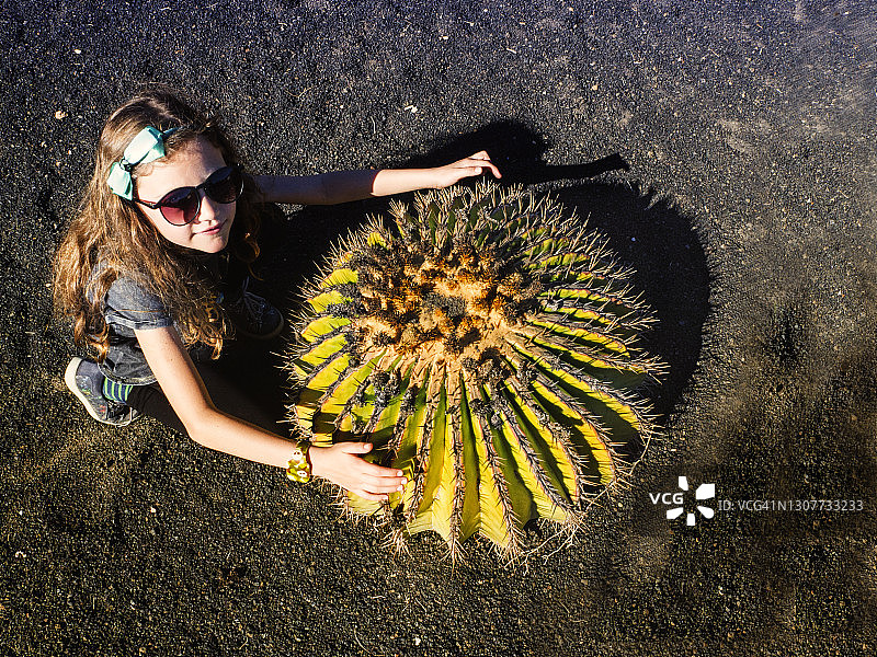 微笑的女孩蹲在一个大圆形仙人掌旁，兰萨罗特，加那利群岛，西班牙图片素材