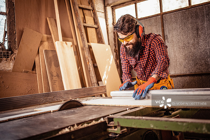 微笑的年轻男子在木工用圆锯砍柴。木工从事木材工艺生产建筑材料或木制家具的木匠。图片素材