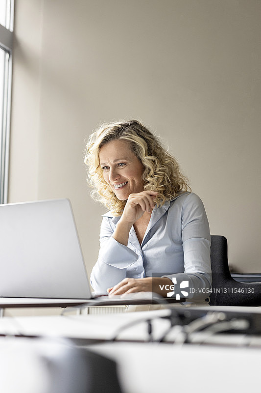 微笑的女企业家坐在办公室看着笔记本电脑图片素材