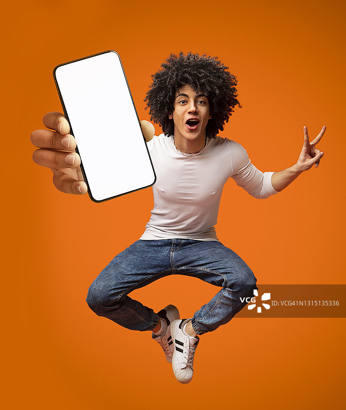 兴奋的非裔美国少年拿着智能手机跳起来，在橙色背景上展示空屏幕图片素材
