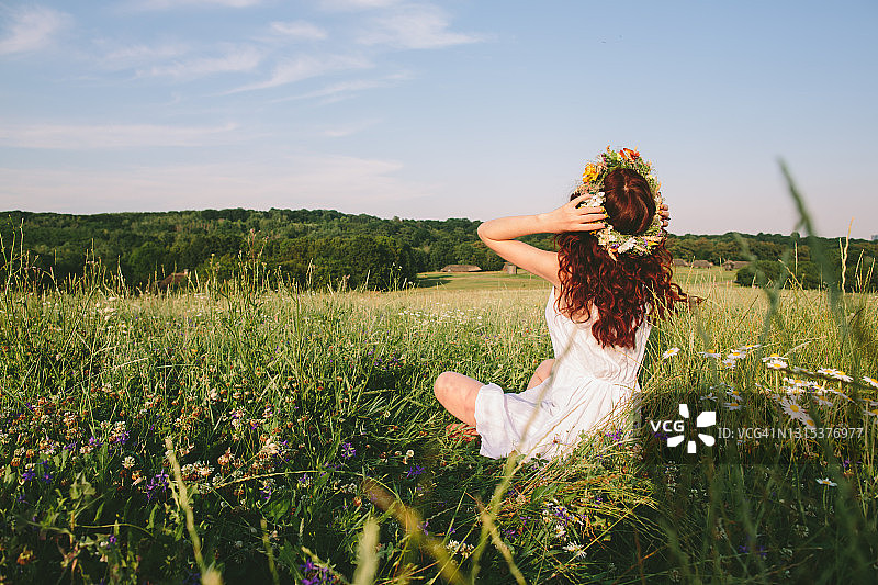 年轻的红发女子戴着春天的花环，坐在草地上休息，微笑，思考，享受夏天的温暖和她的生活图片素材