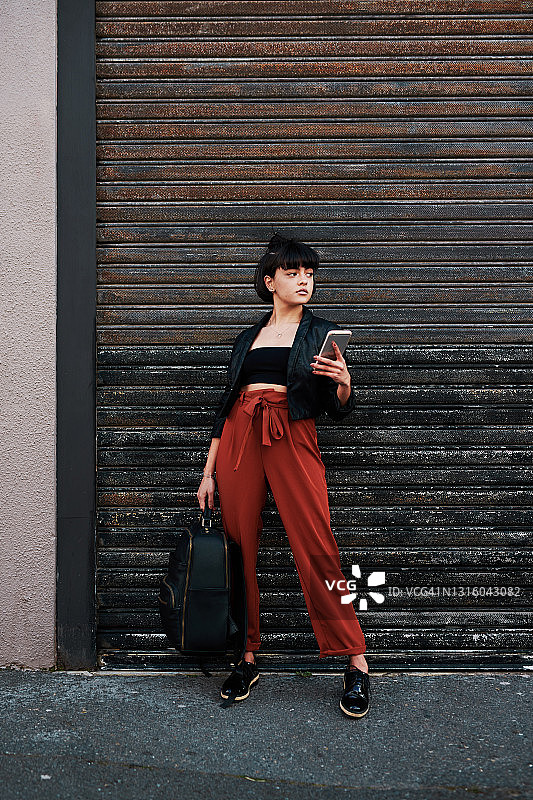 照片中，一名年轻女子拿着背包用手机站在城市的一间车库门前图片素材