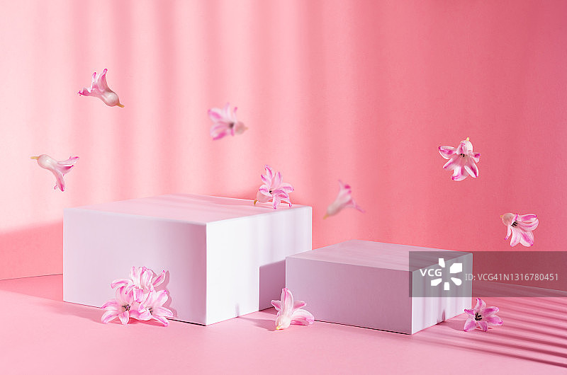 两套白色的方形讲台，展示化妆品和生产在阳光与条纹阴影与春天的花朵飞在柔和的淡粉色背景。图片素材