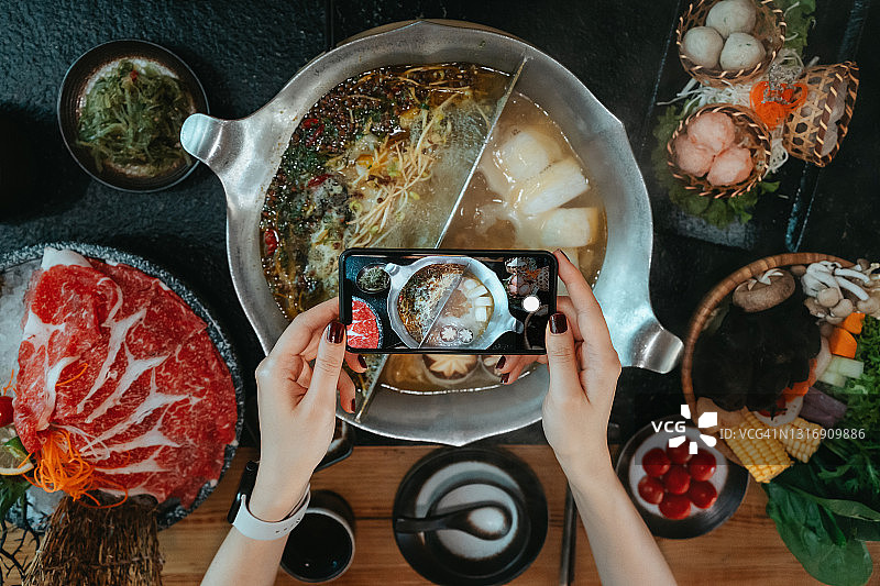 在餐厅吃火锅之前，一名女性用智能手机拍下了美味的中国传统火锅的照片，并分享到社交媒体上。中国菜。外出就餐的生活方式。相机先吃文化图片素材