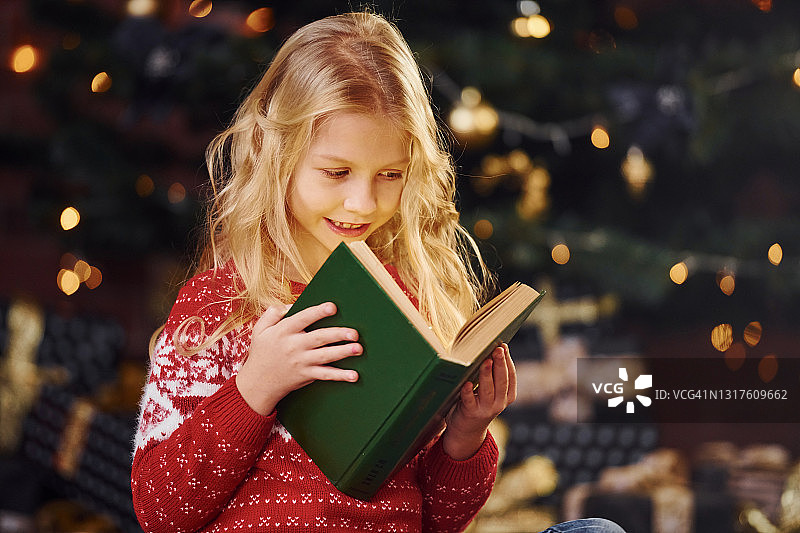 可爱的小女孩在圣诞节期间穿着红色的节日毛衣在室内看书图片素材
