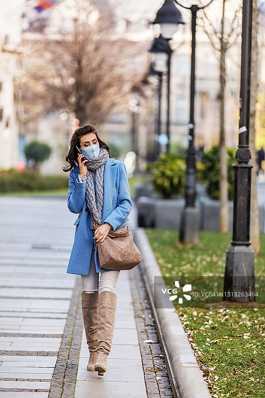 一个戴着面具的漂亮女人在城市的街道上走着，并且在用电话。图片素材