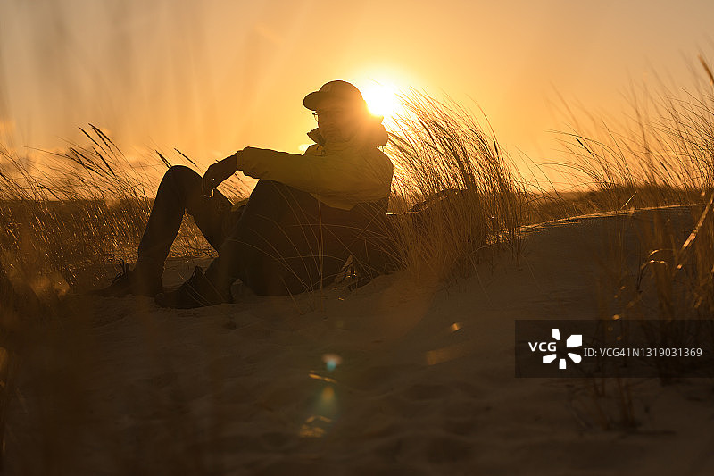 秋天的海滩放松。看着大海日落的人。坐在芦苇间的沙丘上图片素材