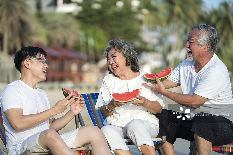 多代幸福家庭坐在沙滩上吃西瓜。他们边吃边笑。图片素材