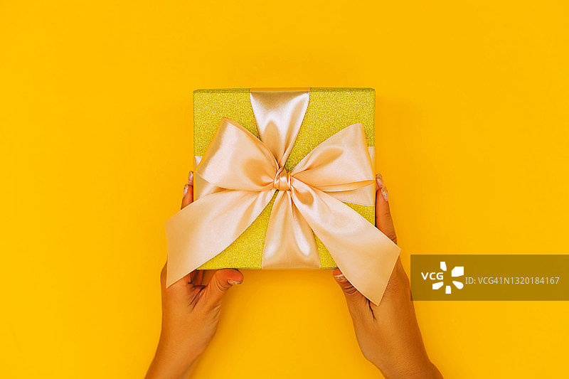 女人的手指甲握着金色的礼品盒与丝绸米色蝴蝶结在明亮的黄色背景。2021年的流行颜色。圣诞节庆祝活动的概念。平的风格。为你的设计复制空间图片素材