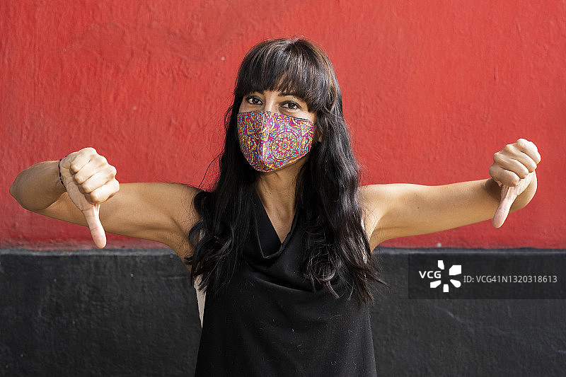 拉丁裔妇女戴着面具，看着摄像机，给两个拇指向下，红色和黑色的背景图片素材
