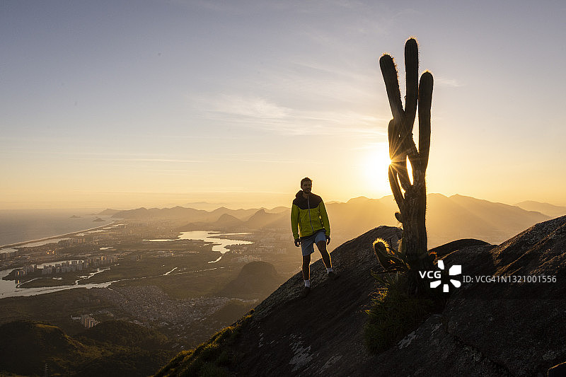 夕阳下，一名男子站在岩石山的边缘，旁边是一棵大仙人掌图片素材