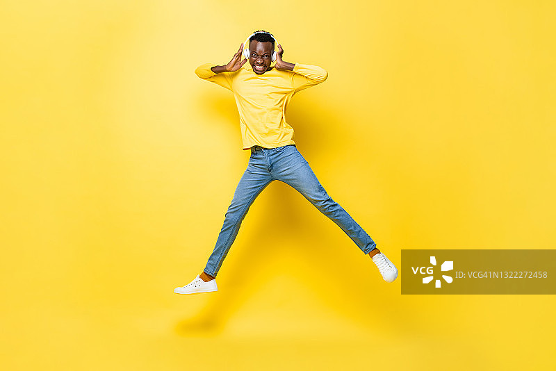 快乐的年轻非洲人戴着耳机听音乐和跳跃在黄色的孤立的演播室背景图片素材