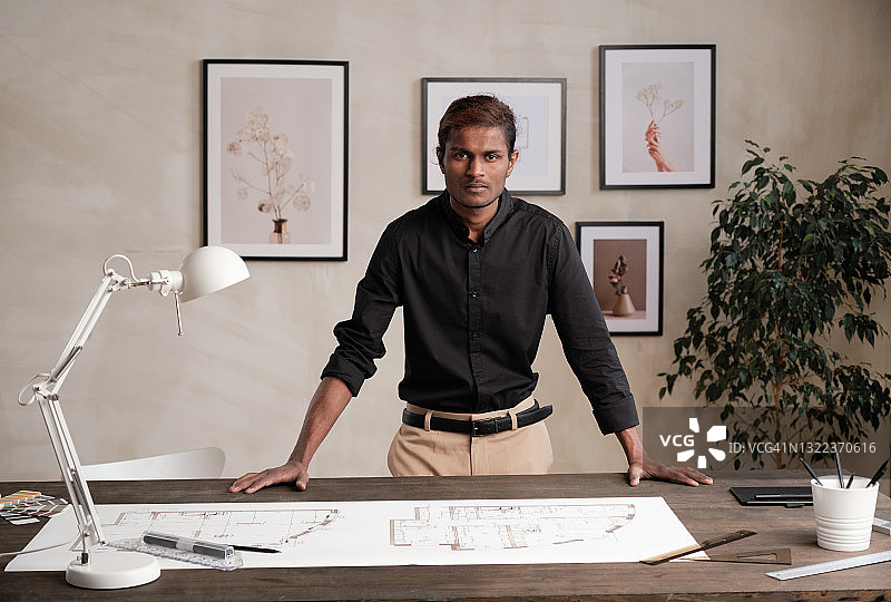 肖像严肃的年轻黑人工程师站在桌子与蓝图和用品在现代办公室墙上的图片图片素材