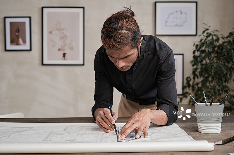 忙碌的年轻混合种族工程师在黑衬衫站在桌子上，靠在它，而使用尺子和图纸上的平面图线图片素材