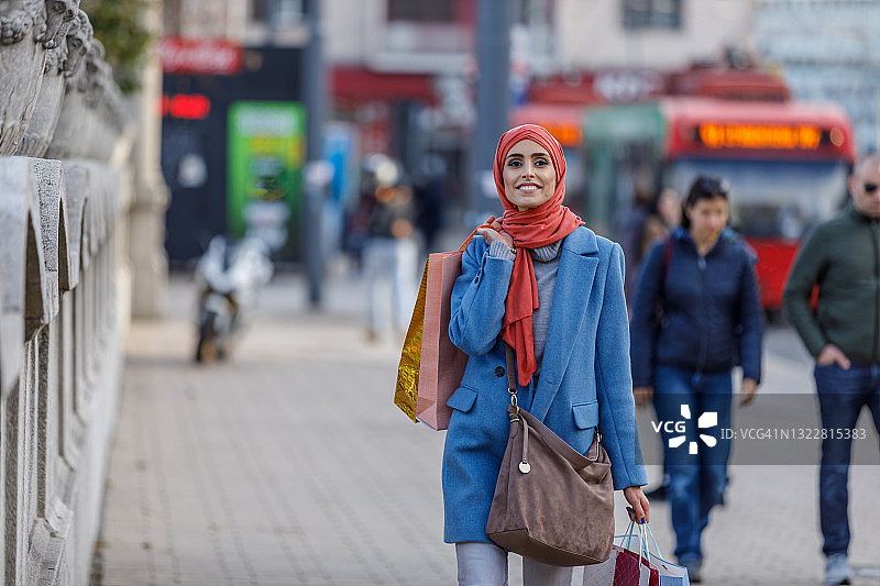 一名兴高采烈的穆斯林妇女的肖像，她拿着购物袋走过城市的街道。图片素材