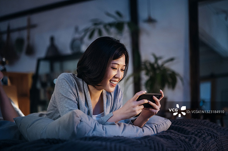 一个年轻的亚洲女人躺在卧室的床上，晚上在舒适的家里用智能手机玩手机游戏图片素材