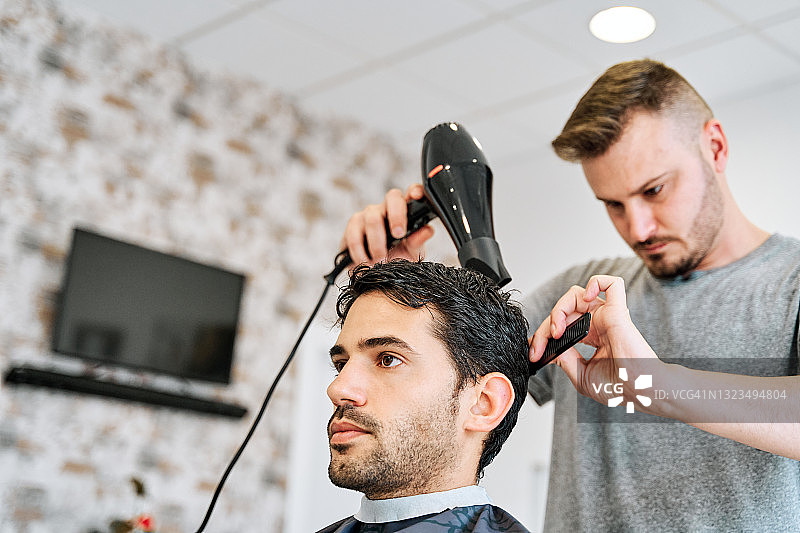 一个理发师在理发店用吹风机吹干一个年轻人的头发的侧面视图。图片素材