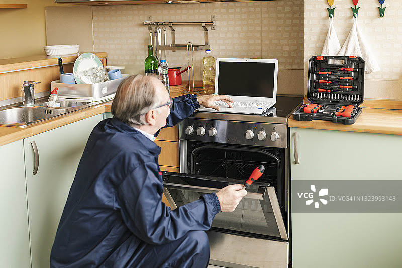 高级工人正在用螺丝刀修理烤箱并寻求在线帮助。图片素材