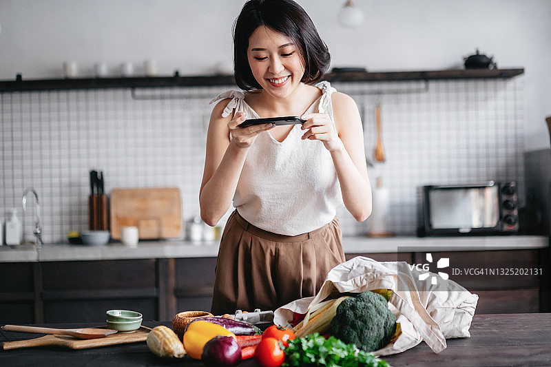 年轻的亚洲妇女从杂货店购物回来，从厨房柜台上的可重复使用的购物袋里拿出新鲜的水果和蔬菜。她正在家里用智能手机给新鲜的农产品拍照图片素材
