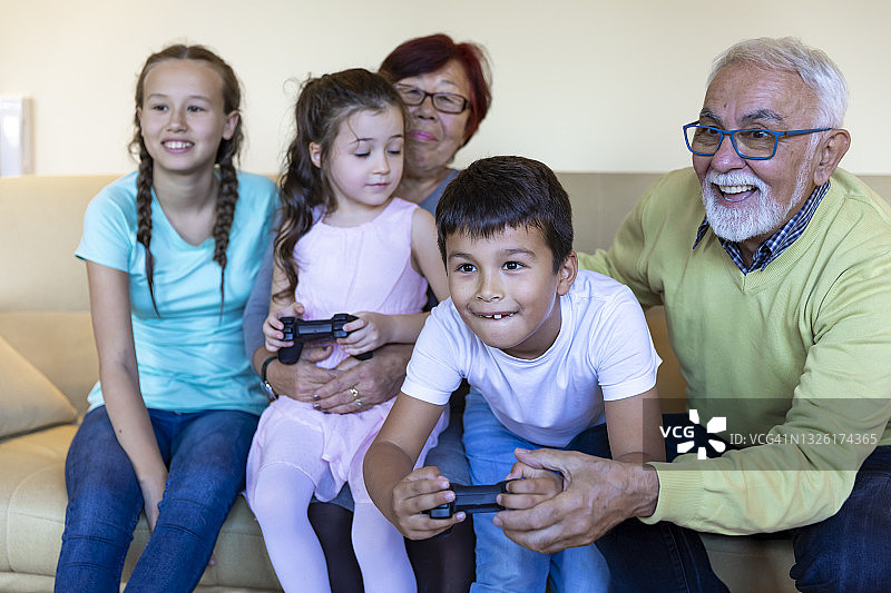 微笑的孩子坐在客厅的沙发上，与大家庭一起享受时光，同时玩主机游戏。图片素材
