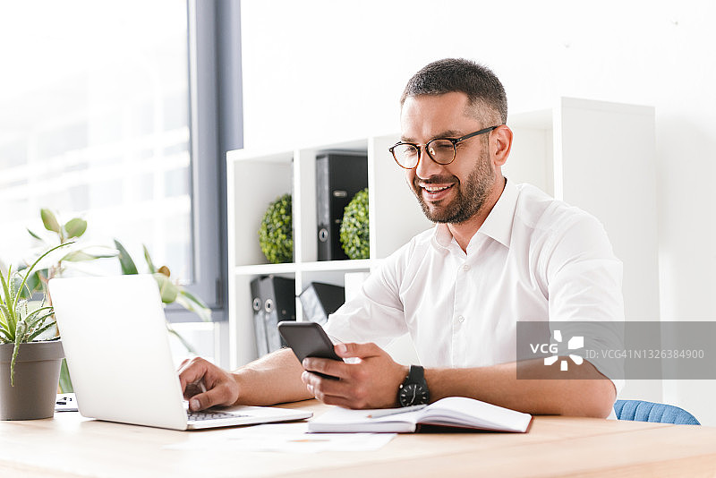 照片中的30多岁的成年男子穿着白衬衫，坐在办公室里使用笔记本电脑和手机图片素材