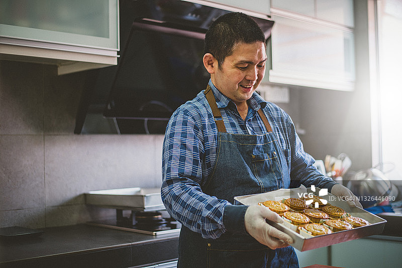 一名亚洲男子从烤箱烤好月饼后，拿着自制的月饼放在烤盘上图片素材