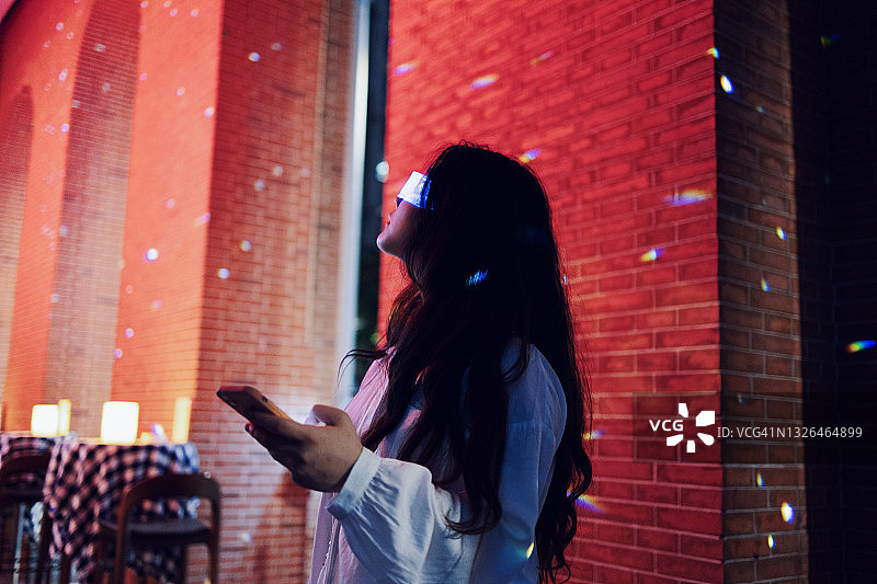 一名女子戴着增强现实眼镜，用智能手机站在夜间的街道上图片素材