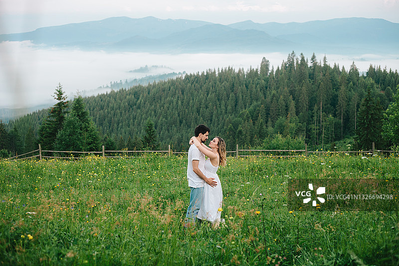 户外浪漫的年轻夫妇肖像。美丽的夏季景观，山与雾，山丘，森林和黄色的野花绿色草地。图片素材