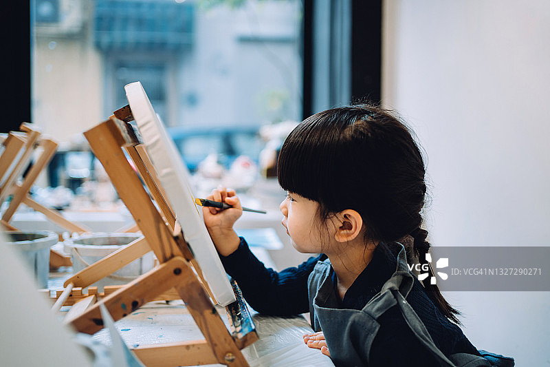 在绘画课上，一个专心致志的亚洲小女孩用画笔和五颜六色的丙烯颜料在画布上作画图片素材