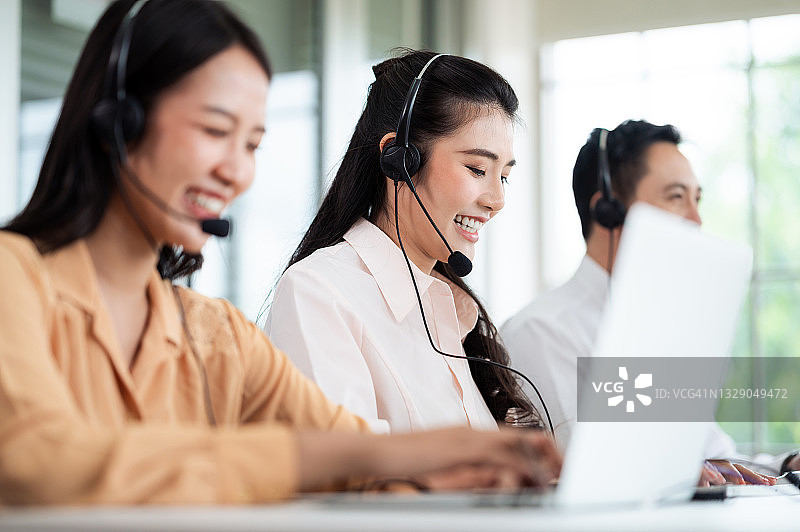 亚洲呼叫中心的工作人员戴着带有麦克风的耳机。在办公桌和电脑前服务顾客时微笑。服务理念与咨询。通信的概念。图片素材