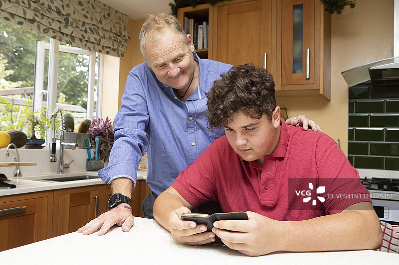 成熟的男人和十几岁的男孩一起看手机在一个家庭厨房图片素材