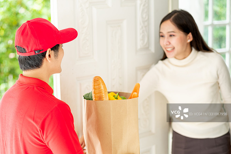身穿红色制服的亚洲送餐员微笑着给开门取货的女士送餐袋图片素材