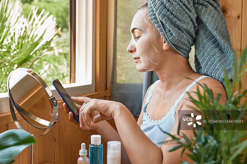 护肤习惯。中年妇女用保湿面膜在手机上上网。提升眼下抗皱面膜。胶原蛋白面膜，护肤和水疗概念。图片素材