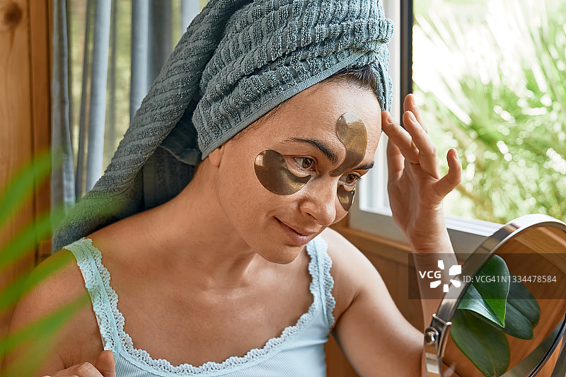 护肤习惯。中年妇女摸着脸上的皱纹和水凝胶眼罩看着镜子。提升眼下抗皱面膜。胶原蛋白面膜，护肤和水疗概念。图片素材