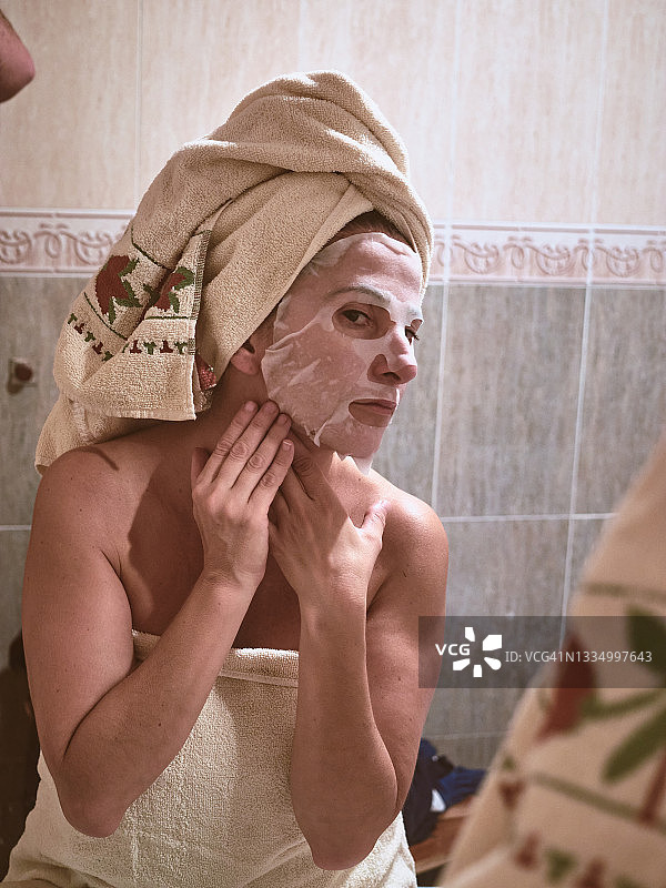 女人早上沐浴后照在镜子前，用毛巾敷在头上敷一张面膜，预防早期的皱纹，护肤时使用润肤霜，美容护肤，护肤理念图片素材