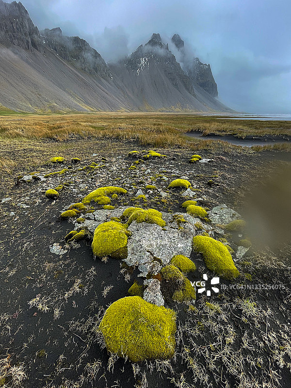 POV的人看着苔藓覆盖的岩石在潮坪与戏剧性的山脉笼罩在雾风暴云。图片素材