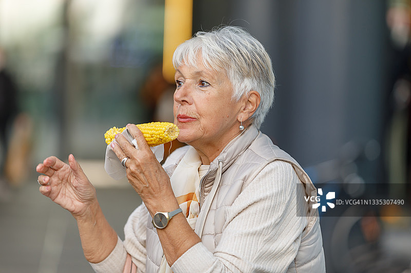 一位老妇人在城市里散步，吃着玉米。图片素材