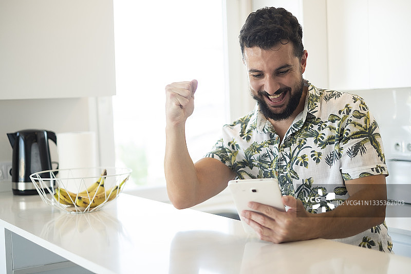 一名兴高采烈的男子在家里的厨房里看着平板电脑，在网上庆祝胜利或收获图片素材