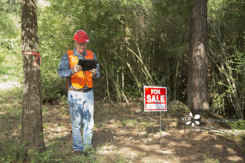 施工经理正在标记要砍伐的树木。他随身携带一台电子平板电脑，穿着安全背心，戴着安全帽。房屋上挂着出售标志。图片素材